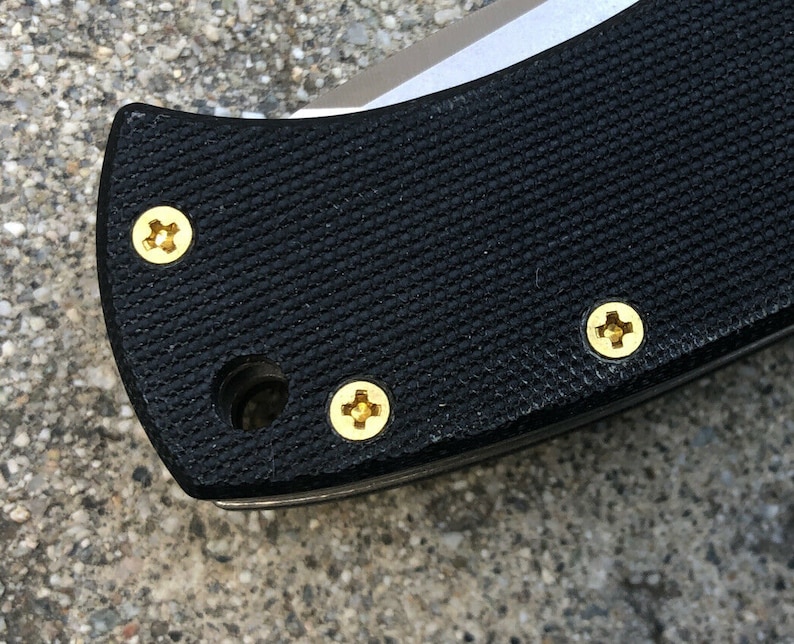 Puede incluir: Primer plano de un mango de cuchillo negro con cuatro tornillos dorados.