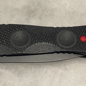 May include: Black folding knife with a textured handle and red accents. The handle has a textured pattern and three circular indentations.