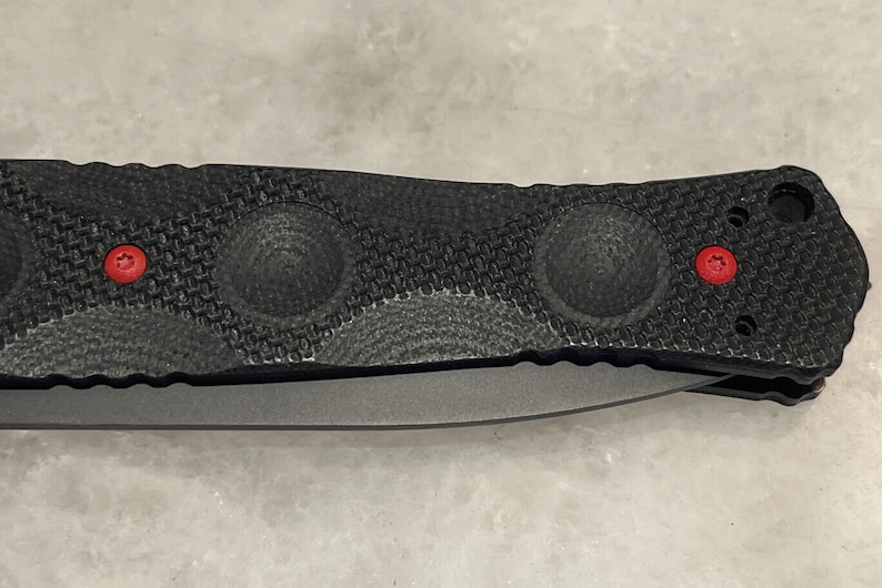 May include: Black folding knife with a textured handle and red accents. The handle has a textured pattern and three circular indentations.