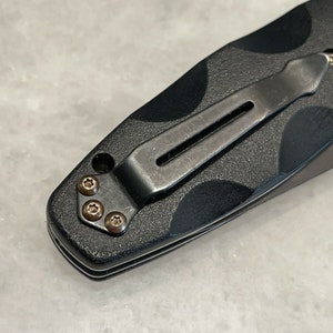May include: Close-up of a black pocket knife with a clip and four screws. The knife has a textured black handle.