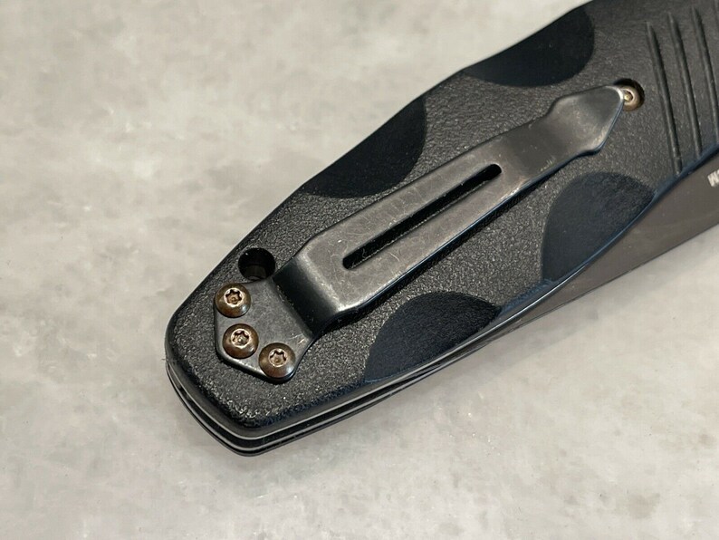 May include: Close-up of a black pocket knife with a clip and four screws. The knife has a textured black handle.