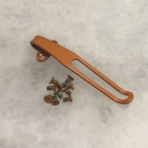 Puede incluir: Un clip de bolsillo de metal naranja con un bucle en el extremo. Hay varios tornillos pequeños al lado del clip.