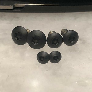 May include: A set of seven black screws with a star-shaped head. The screws are arranged in a semi-circle on a white surface.