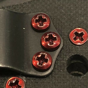 May include: Five red screws with a cross-shaped head are attached to a black surface. The screws are shiny and have a metallic finish.