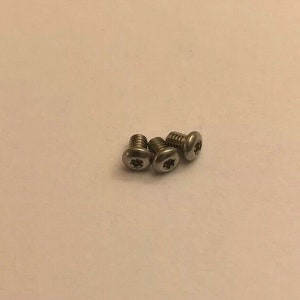 Puede incluir: Tres tornillos pequeños de color plateado con una hendidura en forma de estrella en la cabeza. Los tornillos están dispuestos en un pequeño grupo sobre un fondo liso blanquecino. Los tornillos parecen ser de metal.