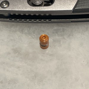 May include: A small, copper-colored, cylindrical object with a textured surface and a small hole in the center.