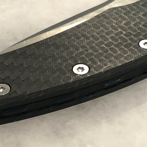 以下が含まれることがあります： 黒いカーボンファイバー製のナイフのハンドルを銀色のネジで留めたクローズアップ。