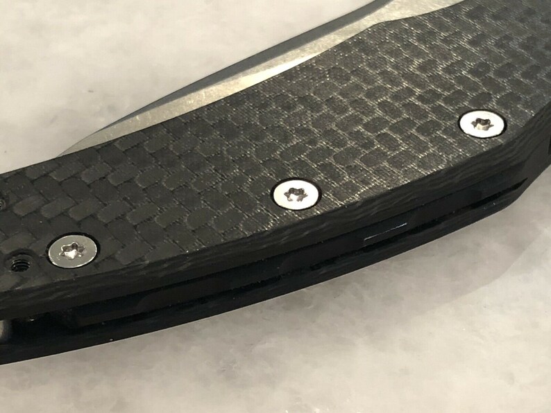 以下が含まれることがあります： 黒いカーボンファイバー製のナイフのハンドルを銀色のネジで留めたクローズアップ。