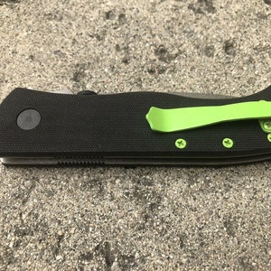 Pode incluir: Uma faca dobr&aacute;vel preta com um clipe de bolso verde e uma al&ccedil;a preta texturizada. A faca tem uma l&acirc;mina prateada e v&aacute;rios parafusos.
