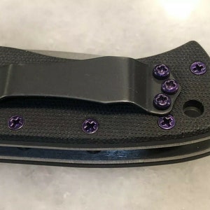 May include: A black pocket knife with purple screw detail. The knife has a clip on the back and a textured handle.