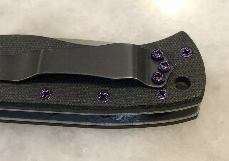 May include: A black pocket knife with purple screw detail. The knife has a clip on the back and a textured handle.