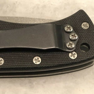 May include: Close-up of a black pocket knife with a textured handle and a silver blade. The knife has a pocket clip and several screws on the handle.