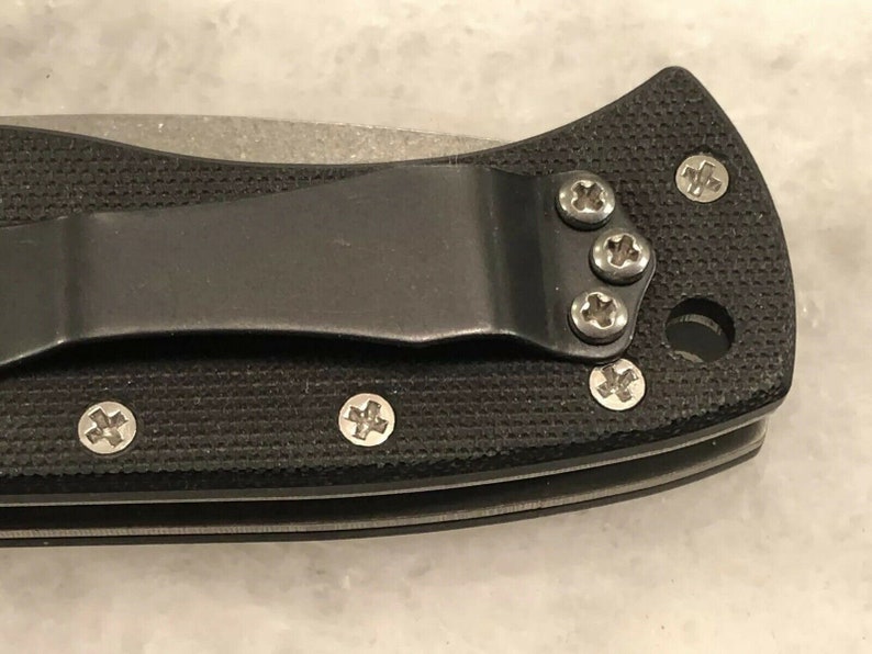 May include: Close-up of a black pocket knife with a textured handle and a silver blade. The knife has a pocket clip and several screws on the handle.