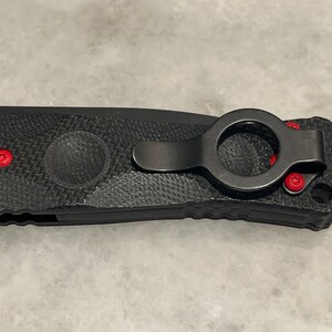 May include: Black folding knife with a textured handle and a black pocket clip with a red ring. The knife has red accents on the handle.