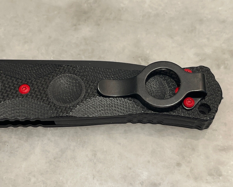 May include: Black folding knife with a textured handle and a black pocket clip with a red ring. The knife has red accents on the handle.