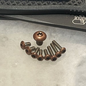 May include: A collection of bronze-colored screws of varying sizes arranged in a semi-circle. The screws have a star-shaped recess in their heads. A larger, domed screw sits in the center. A black knife is in the background.
