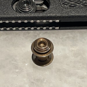 May include: A close-up of a small, round, brass-colored button with a raised center and a smooth, flat bottom.