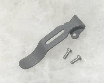 May include: A gray metal pocket clip with two silver screws. The clip is designed to attach to a pocket or belt.