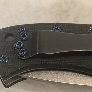 May include: Close-up of a black pocket knife with a clip and blue screws. The knife has a textured handle and a silver blade.