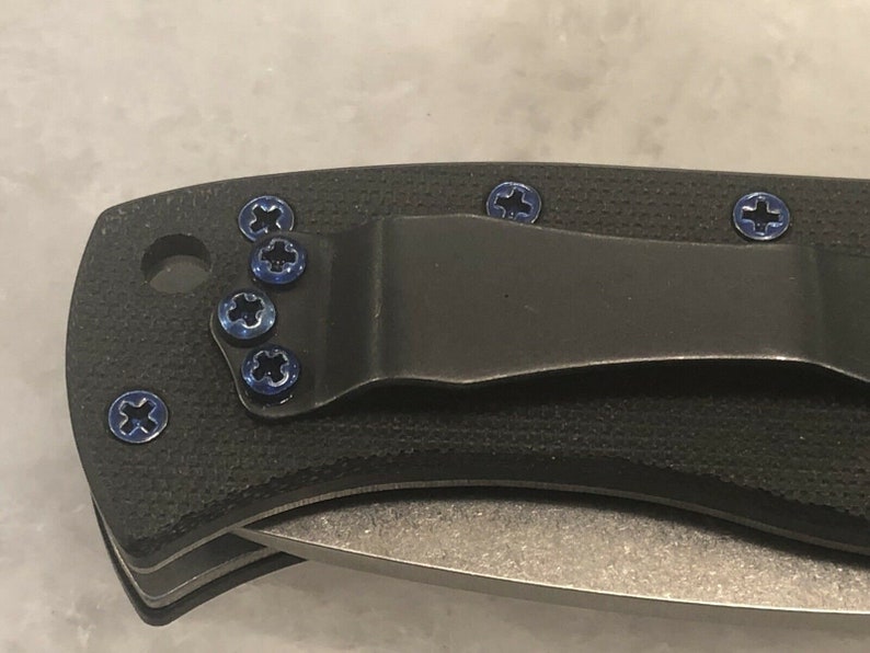 May include: Close-up of a black pocket knife with a clip and blue screws. The knife has a textured handle and a silver blade.