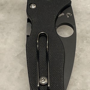 K&ouml;nnte beinhalten: Schwarzes Taschenmesser mit strukturiertem Griff und einem Clip. Das Messer hat ein kleines Loch an der Seite f&uuml;r eine Schnur.