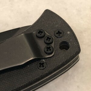 May include: Close-up of a black pocket knife with a clip and four black screws.
