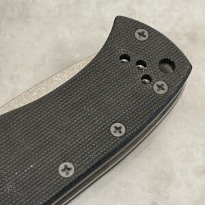 May include: Close-up of a black pocket knife handle with a textured surface and multiple screws. The handle is made of a durable material and has a sleek design.