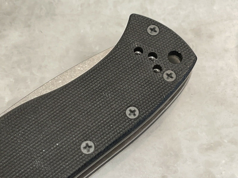 May include: Close-up of a black pocket knife handle with a textured surface and multiple screws. The handle is made of a durable material and has a sleek design.