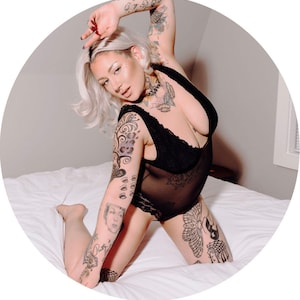 Könnte beinhalten: Eine Frau mit Tattoos posiert auf einem weißen Bett, bekleidet mit einem schwarzen Spitzenbody. Sie hat blonde Haare und schaut in die Kamera.