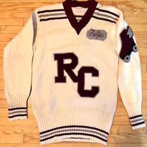 May include: A white vintage sweater with maroon and grey stripes. The sweater has a maroon v-neck and a large maroon and grey "RC" patch on the front. The right sleeve has a maroon patch with a roller skate and wings. The sweater also has a maroon patch with the name "Felipe" on the front.