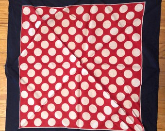 May include: A square scarf with a navy blue border and a red center featuring large white polka dots. The scarf is folded diagonally, creating a unique pattern. The fabric appears to be lightweight.
