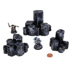 May include: A collection of dark grey miniature rock formations, suitable for tabletop gaming. The image includes two painted figures and a penny for scale. The rocks vary in size and shape.