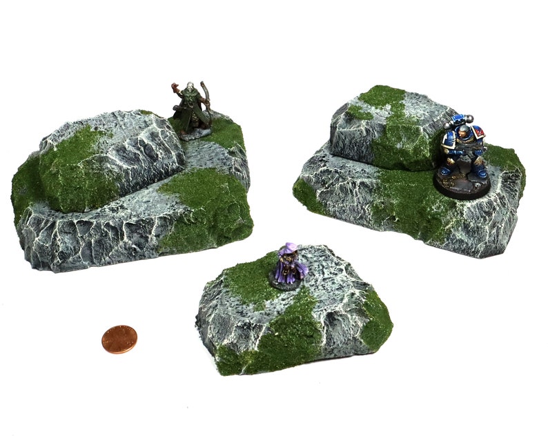 May include: Three gray and green rock formations with a mossy texture. The rocks are different sizes and shapes, and they are perfect for use as terrain in tabletop games.