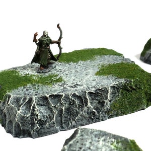 May include: A miniature figure of an elf archer standing on a gray and green rock base. The elf is wearing green clothing and holding a bow and arrow. The base is textured to look like rock and has green moss on the edges.
