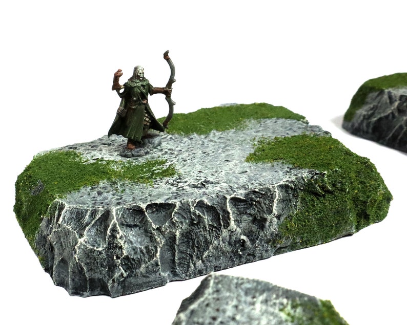 May include: A miniature figure of an elf archer standing on a gray and green rock base. The elf is wearing green clothing and holding a bow and arrow. The base is textured to look like rock and has green moss on the edges.