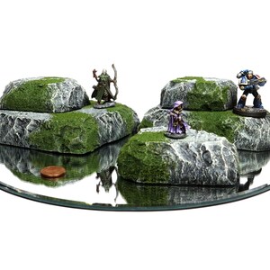 May include: Four miniature rock formations with green moss and gray stone, each with a miniature figure on top. The figures include a green elf with a bow and arrow, a purple-robed figure, and a blue space marine with a gun.