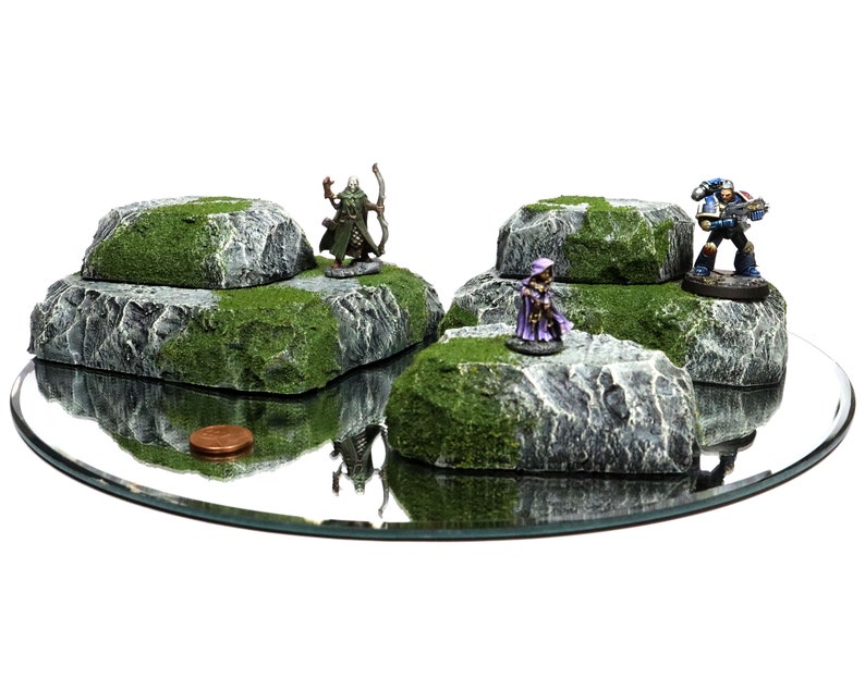 May include: Four miniature rock formations with green moss and gray stone, each with a miniature figure on top. The figures include a green elf with a bow and arrow, a purple-robed figure, and a blue space marine with a gun.