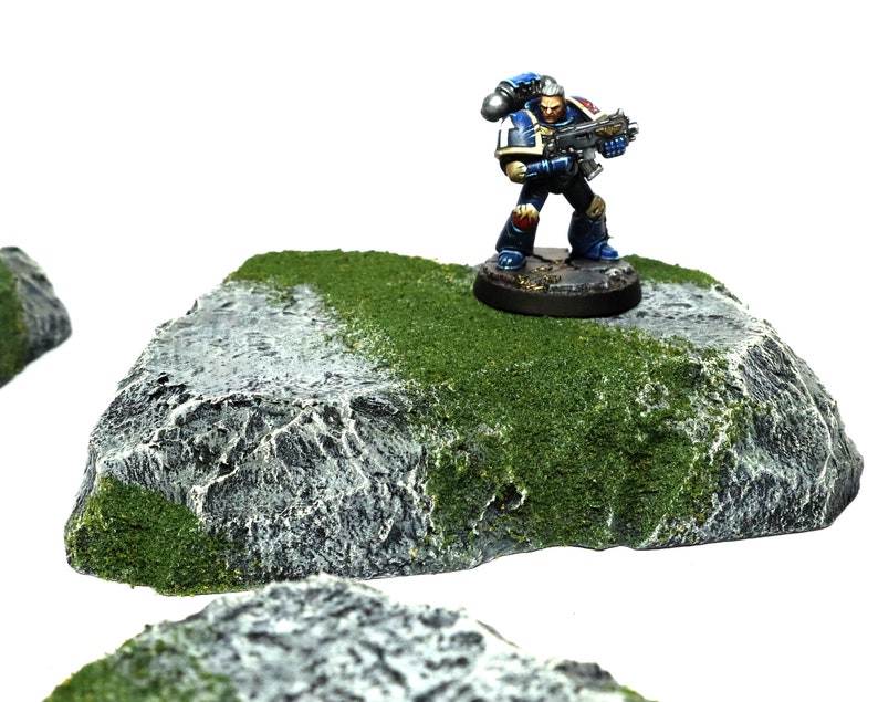 May include: A miniature plastic rock with a green moss texture, perfect for tabletop gaming. A blue and silver painted miniature figure stands on top of the rock.