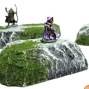 May include: Three miniature rocky terrain pieces with green moss and a miniature figure in a purple robe standing on one of the rocks.