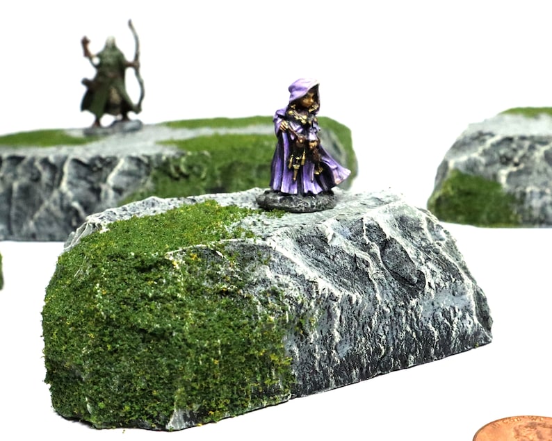 May include: Three miniature rocky terrain pieces with green moss and a miniature figure in a purple robe standing on one of the rocks.