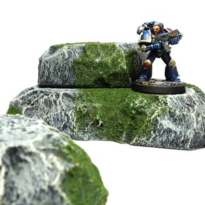 May include: A miniature figure of a space marine standing on a gray rock with green moss. The rock is part of a two-tiered rock formation.