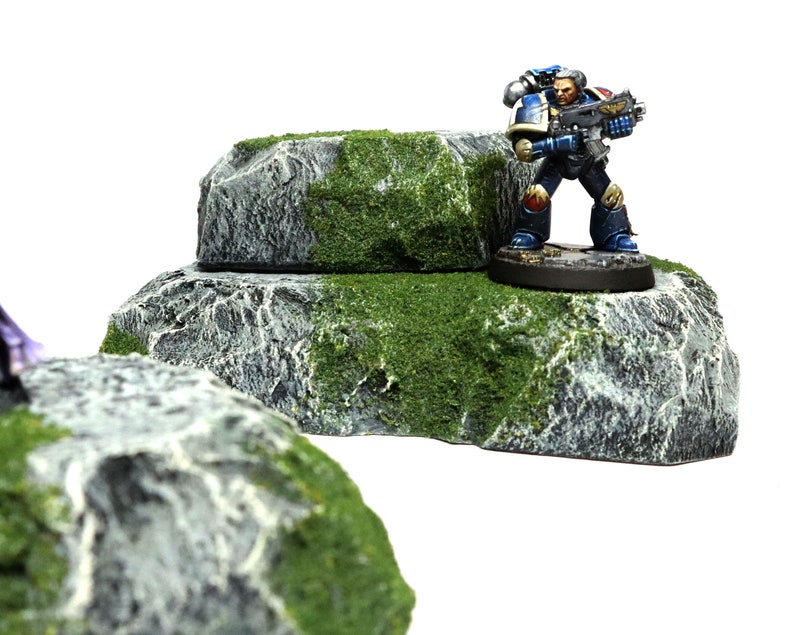 May include: A miniature figure of a space marine standing on a gray rock with green moss. The rock is part of a two-tiered rock formation.