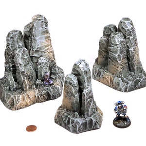 May include: Three grey and brown rock formations, each with a different shape and size. The rock formations are made of a textured material and are designed for use in tabletop games.