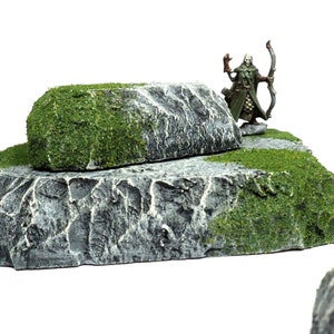 May include: A miniature figure of an elf archer standing on a two-tiered rock formation. The rock is painted gray with green moss accents.