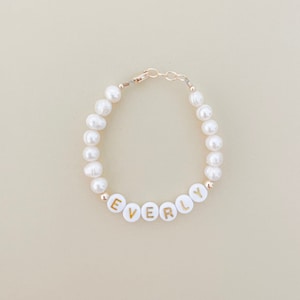 Puede incluir: Un brazalete de perlas blancas con un cierre dorado y cuentas blancas con letras doradas que deletrean "EVERLY".