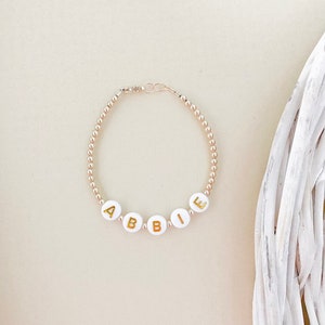 May include: Gold bead bracelet with white letter beads spelling out the name "Abbie".