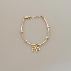 May include: Gold-colored beaded bracelet with a bow charm. The bracelet features small, round beads and larger accent beads. The bow charm has a small, clear gemstone in the center. The bracelet is approximately 7 inches long.