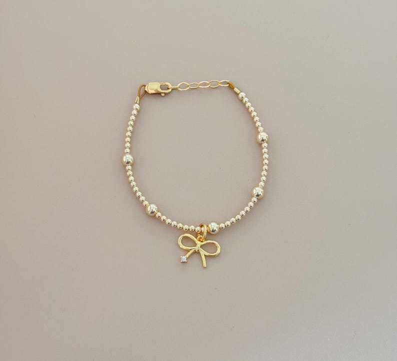 May include: Gold-colored beaded bracelet with a bow charm. The bracelet features small, round beads and larger accent beads. The bow charm has a small, clear gemstone in the center. The bracelet is approximately 7 inches long.