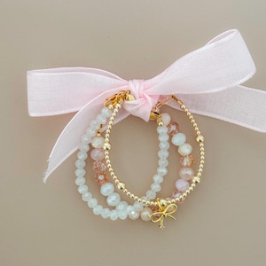 May include: Three beaded bracelets in white, gold, and pink are tied together with a pink ribbon. The bracelets feature small, round gold beads and larger, faceted beads in white and pink. A gold bow charm hangs from one bracelet.