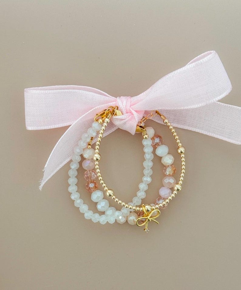 May include: Three beaded bracelets in white, gold, and pink are tied together with a pink ribbon. The bracelets feature small, round gold beads and larger, faceted beads in white and pink. A gold bow charm hangs from one bracelet.